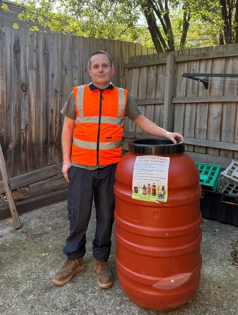 Photo of Alex wearing a hi-vis vest and standing next to a 10c recycling barrel.